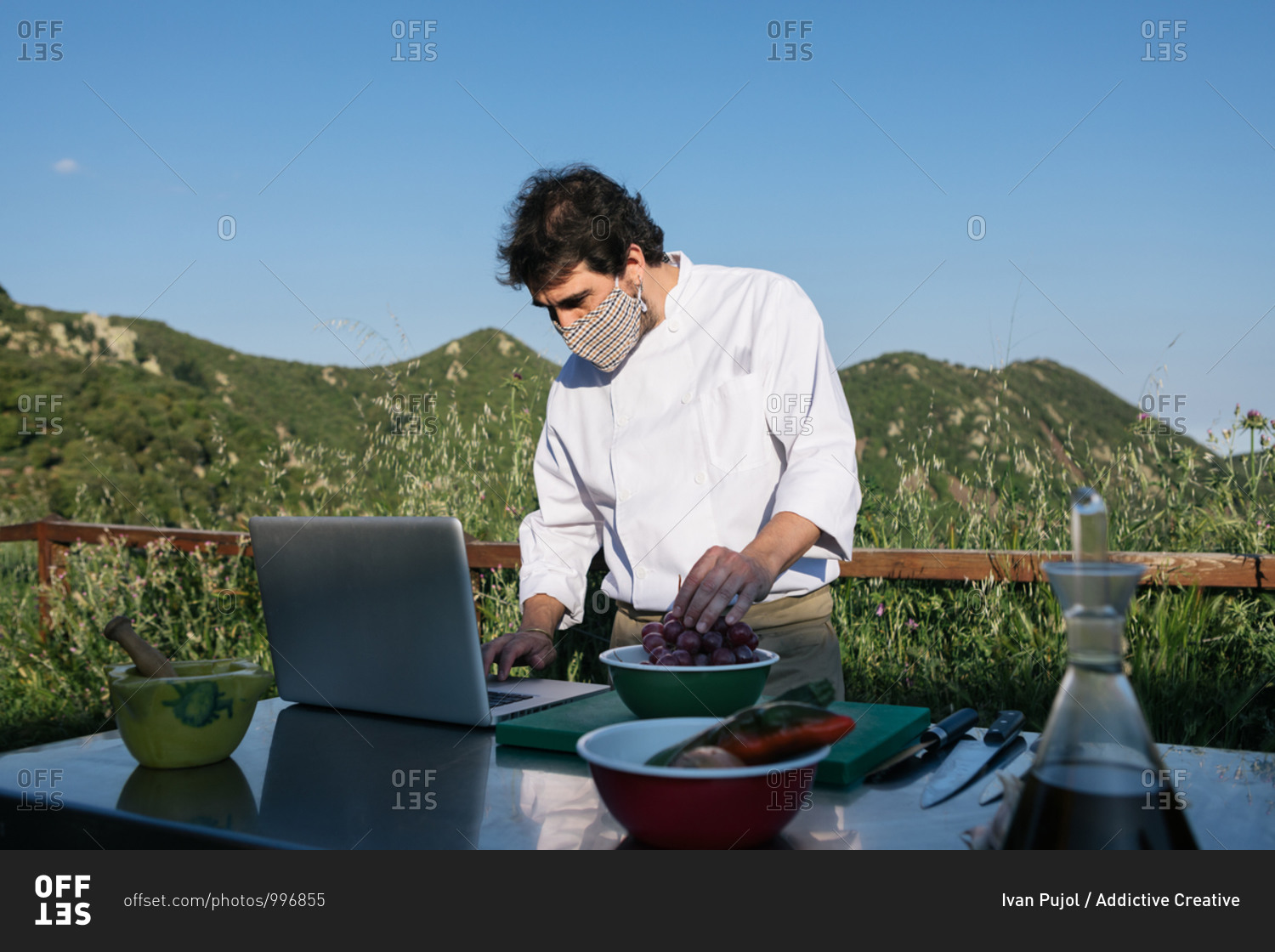 Unrecognizable male chef in uniform and mask checking recipe on laptop while preparing traditional dish lunch in outdoor kitchen during training in countryside