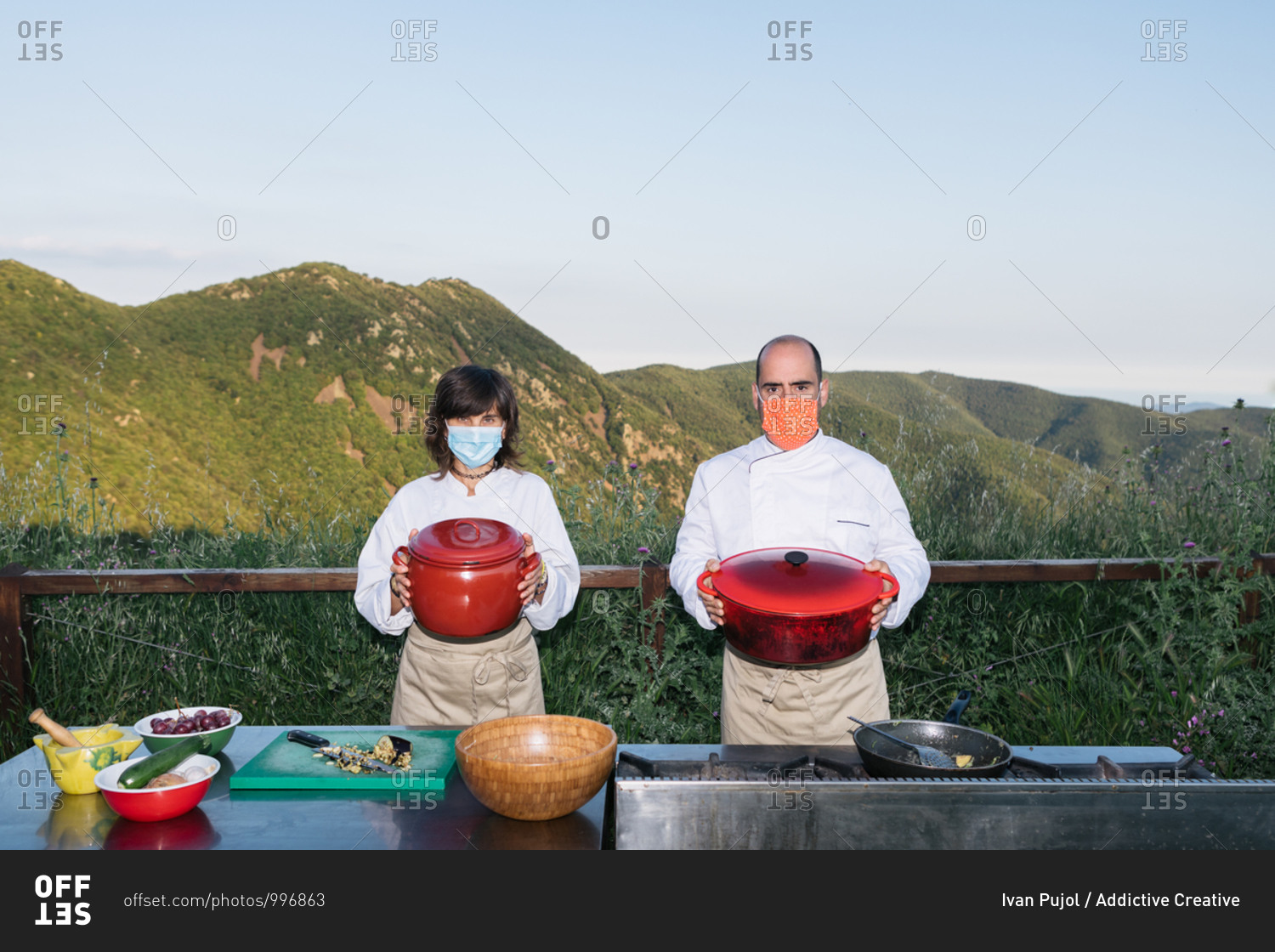 Anonymous professional cooks in uniforms and medical masks standing with saucepans in hands against mountainous terrain on sunny day