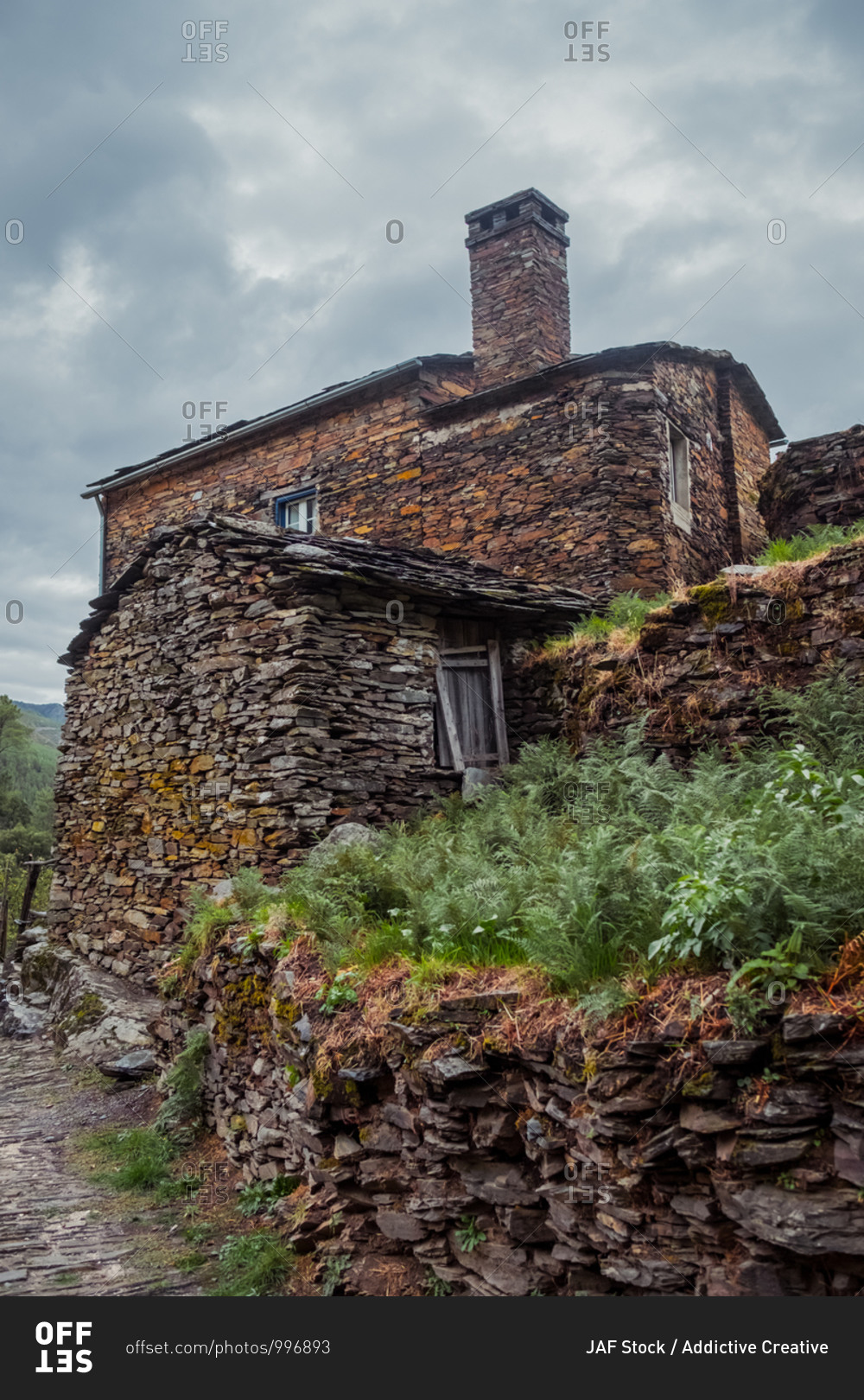 Exterior of old stone house with rough surface and chimney on top near green plants and pathway under cloudy sky