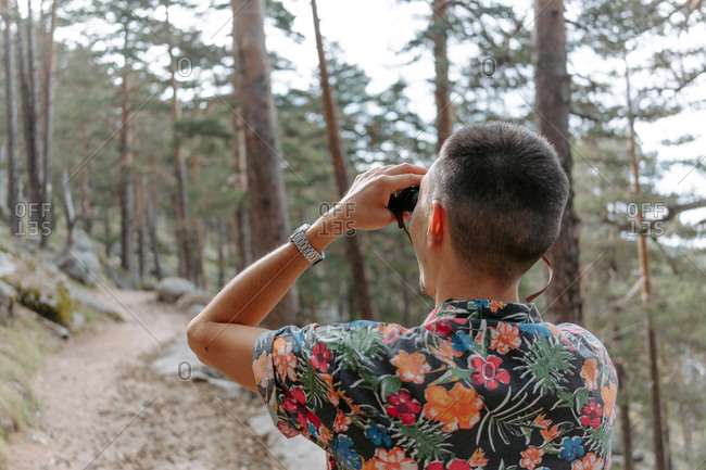 Back view of male tourist in colorful shirt standing on slope of mountain covered with coniferous forest and observing environment with binoculars during hiking in Navacerrada in Spain