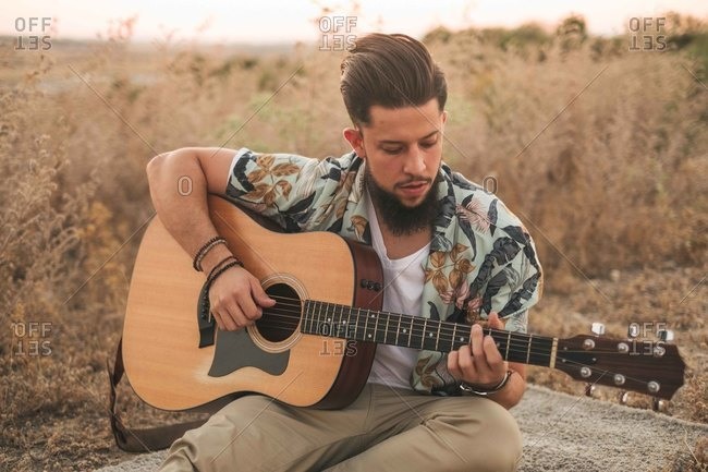 Smiling hipster male musician in casual wear playing classic guitar while sitting on rug near faded grass under serene sky in evening