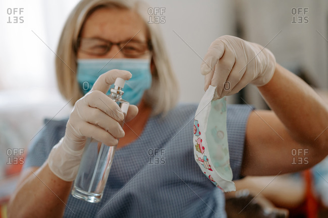Senior female master in protective mask and latex gloves disinfecting handmade fabric mask with sanitizer while producing masks for coronavirus prevention at workplace