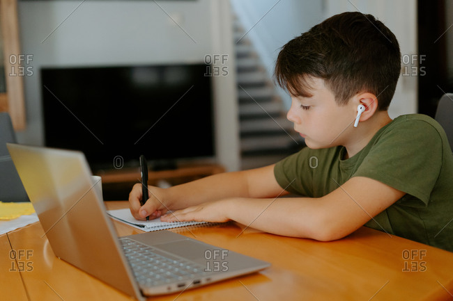 Side view of child writing in spiral notebook while doing homework assignment at table with cup of hot drink