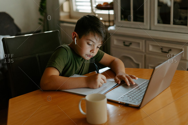 High angle of focused little boy in wireless earphones using laptop and writing down information while doing homework assignment