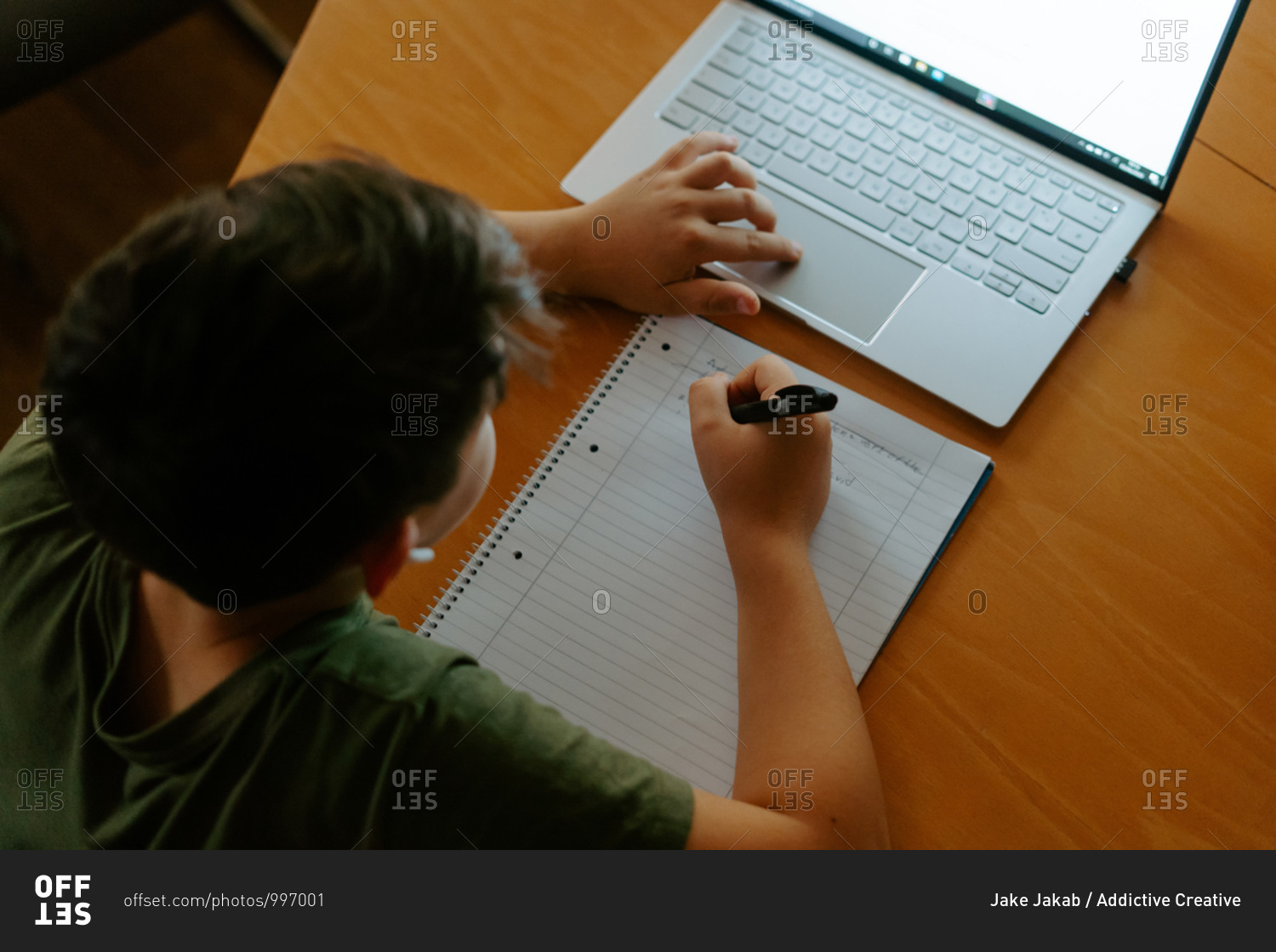 High angle of focused little boy in wireless earphones using laptop and writing down information while doing homework assignment