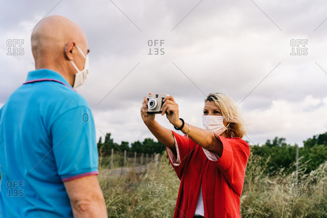 Positive female in face mask taking picture of husband in casual wear using photo camera while spending summer weekend together in countryside during coronavirus epidemic