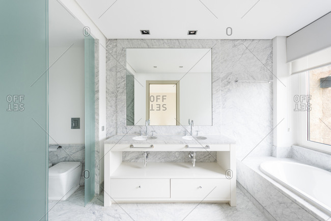 Creative minimalist design of bathroom with similar vanity units under big mirror and bath tube near window