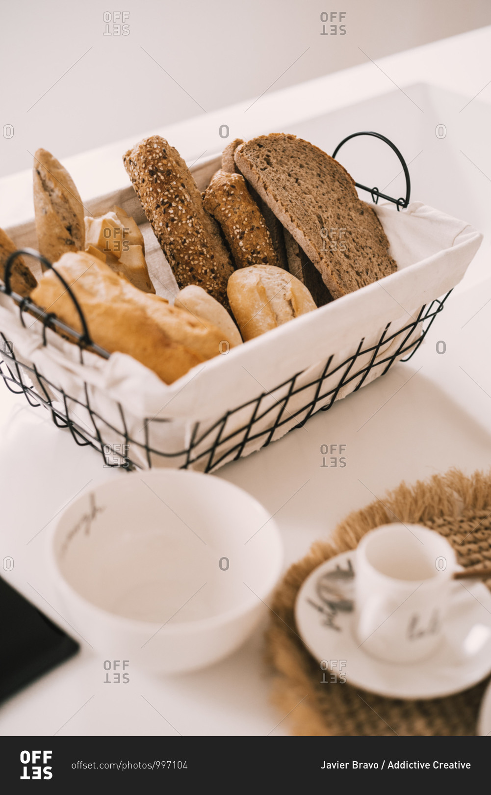 Porcelain tableware set arranged on cloth napkin near basket with fresh bread on table served for breakfast in cozy light apartment