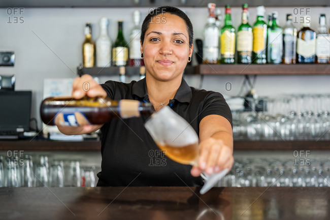 Ethnic positive woman with short hair pouring beer from bottle into glass goblet while working at bar