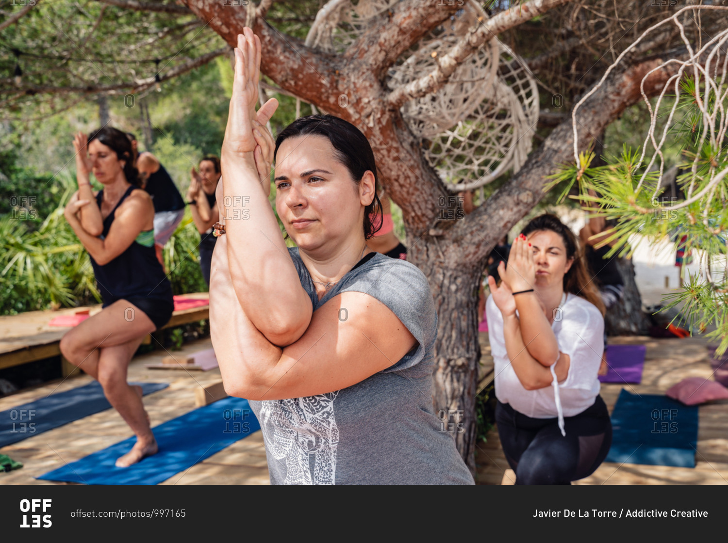 Group of active women in sportswear balancing on leg while practicing Garudasana yoga position during lesson in park on sunny day