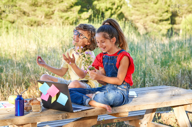 A ten year old boy and girl sitting at a wooden table in the field where there is a computer, flowers, fruit, snack. Dressed in denim and colorful T-shirts.