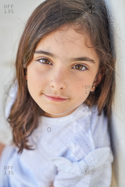 Adorable school aged girl in casual clothes smiling and looking at camera while sitting near wall of house in sunny summer day