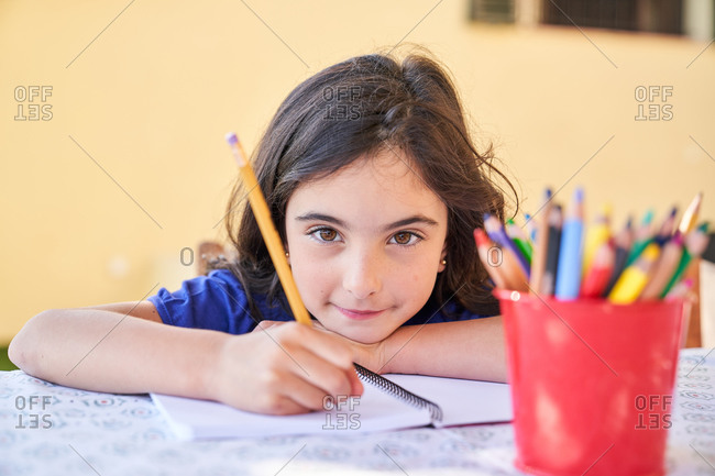 Positive school aged girl looking at camera and smiling while sitting at table and drawing with crayons in copybook during free time