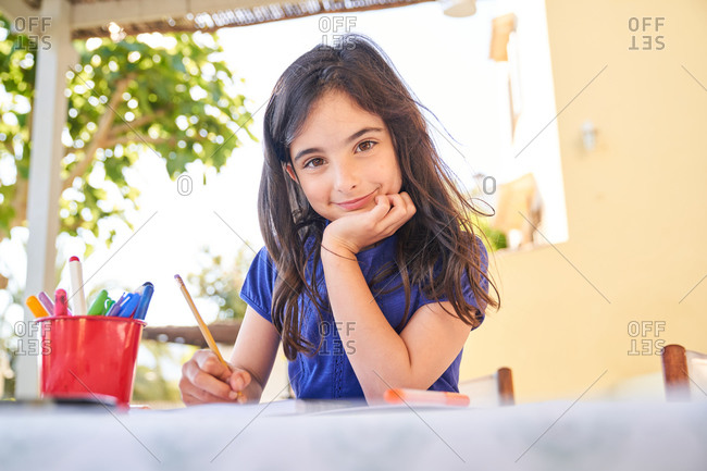 Positive school aged girl while sitting at table and drawing with colorful pencils in copybook during free time