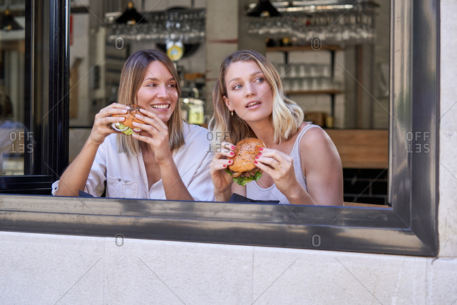 Expressive girlfriends grimacing while sharing burgers in cafe sitting near opened window