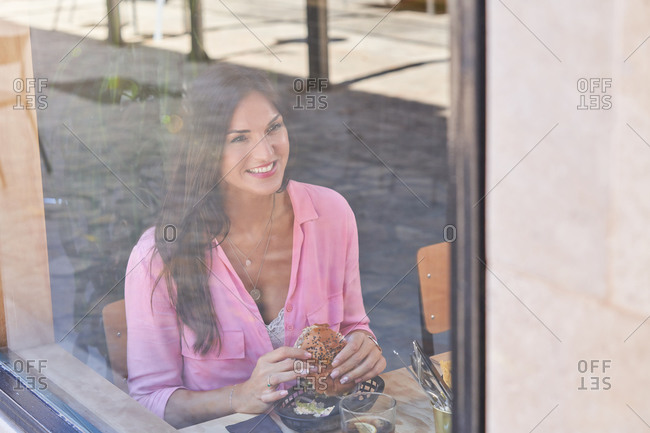 Through window of smiling brunette in pink shirt having delicious burger while smiling away in modern cafe