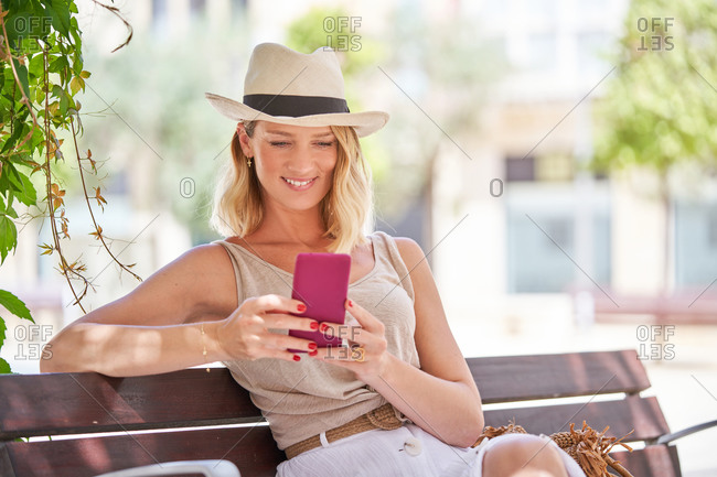 Trendy casual female in hat and skirt browsing smartphone while resting on wooden bench in bright sunny day