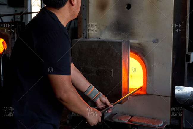 Crop back view of man holding blowpipe and melting glass in hot furnace in workshop