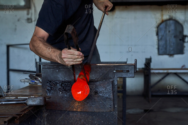 Faceless craftsman heating piece of molten glass on blowpipe while working in manufacture studio