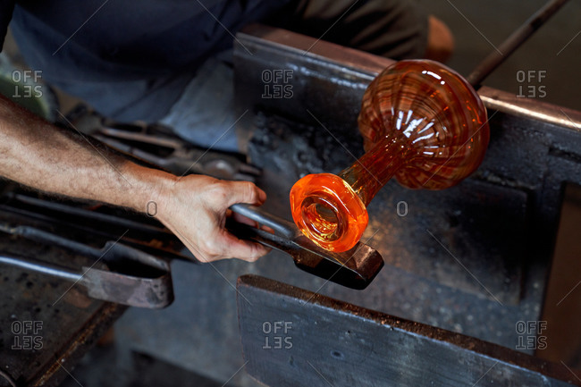 Faceless craftsman heating piece of molten glass on blowpipe while working in manufacture studio