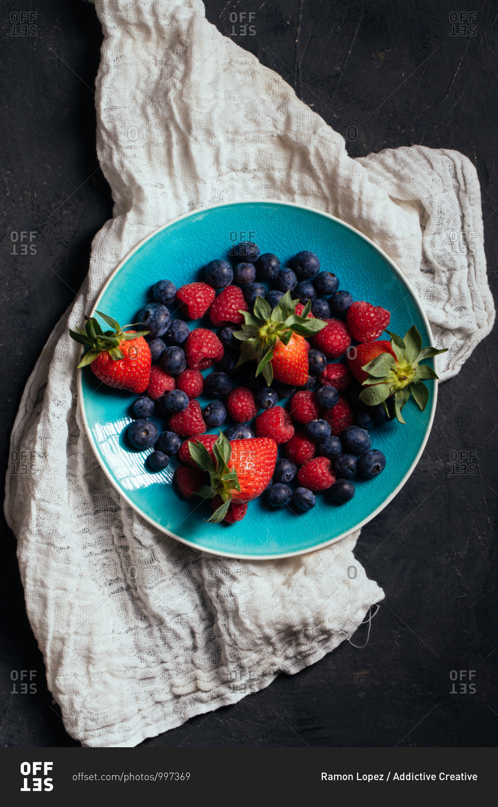 Top view of blue bowl with yummy ripe strawberries and blueberries with raspberries arranged on gray cloth on black background