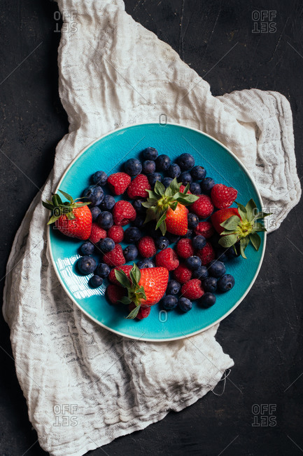 Top view of blue bowl with yummy ripe strawberries and blueberries with raspberries arranged on gray cloth on black background