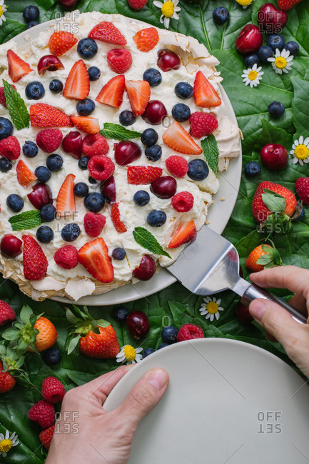 Top view of crop anonymous person with plate and spatula serving delicious summer dessert Pavlova cake with meringue and assorted fresh berries garnished with green leaves