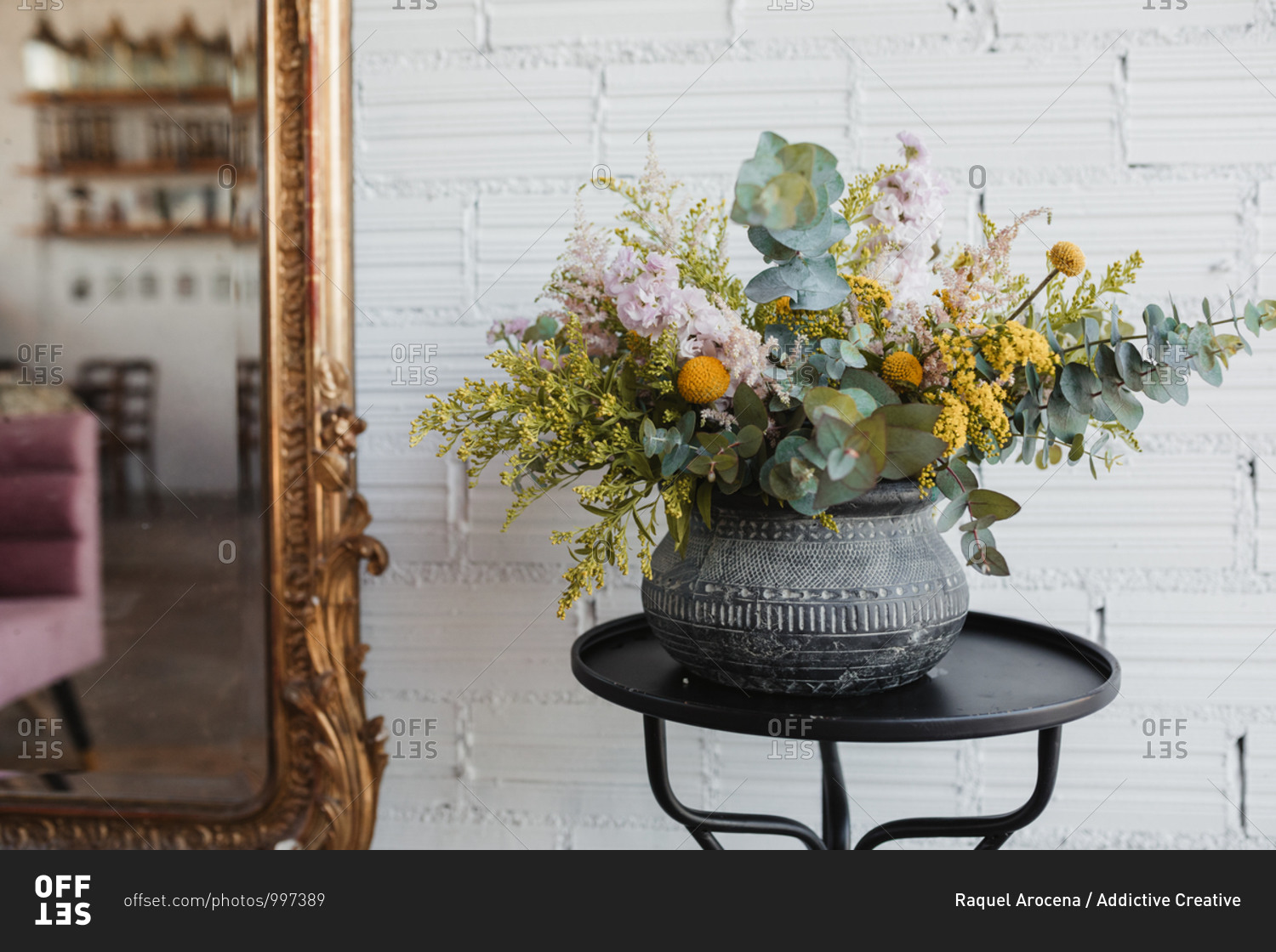 Beautiful bouquet with various flowers including goldenrod and craspedia flowers with green eucalyptus branches arranged in ornamental ceramic pot placed on small table against white brick wall with mirror in creative floristry studio