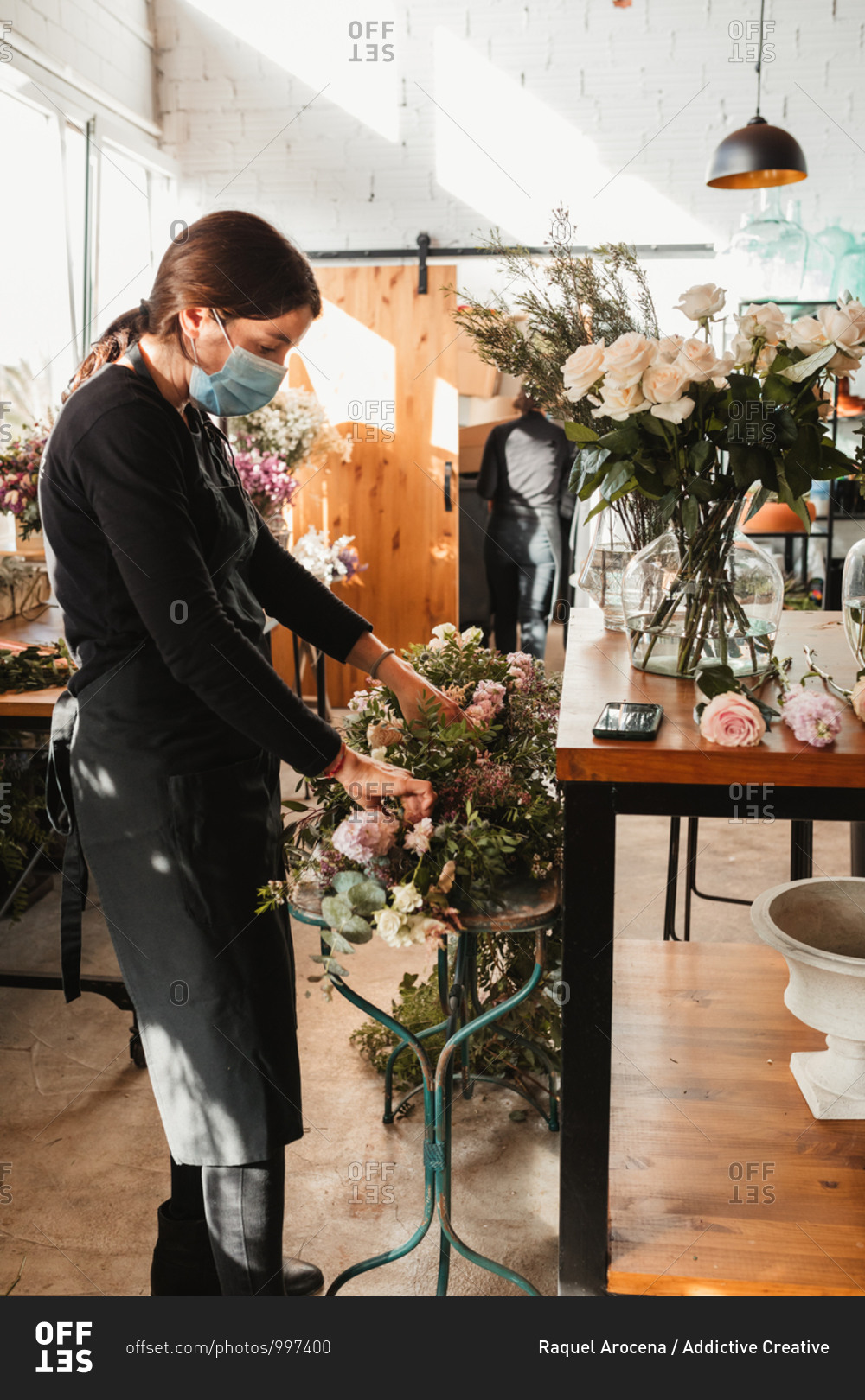 Side view of professional female floral master in protective mask arranging decorative composition with blooming roses while working in floristry workshop