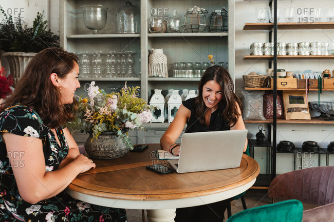 Side view of female owner of stylish floristry salon discussing details of order with customer while sitting together at table with laptop in a happy attitude