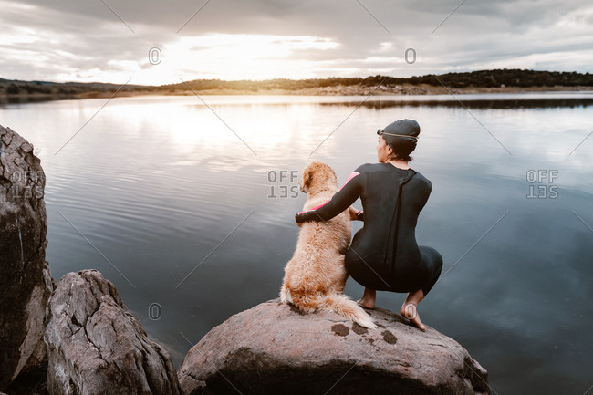 Back view of anonymous woman in black wetsuit standing with dog on stone looking away while observing lake nature