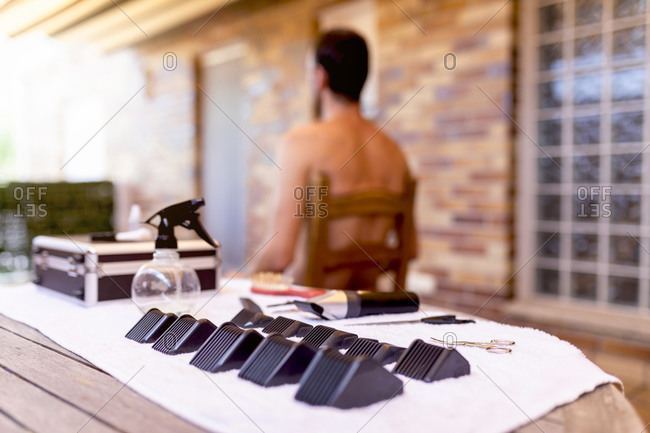 Back view of naked anonymous male sitting on wooden chair near table with kit of plastic trimmer nozzles of different sizes with scissors and trimmer near spray bottle in barbershop