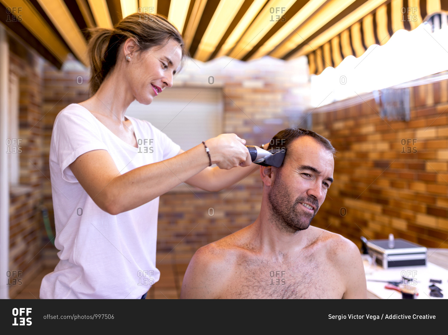 Positive female barber in t shirt using trimmer while doing haircut to shirtless masculine bearded client in hairdressing salon with brick wall