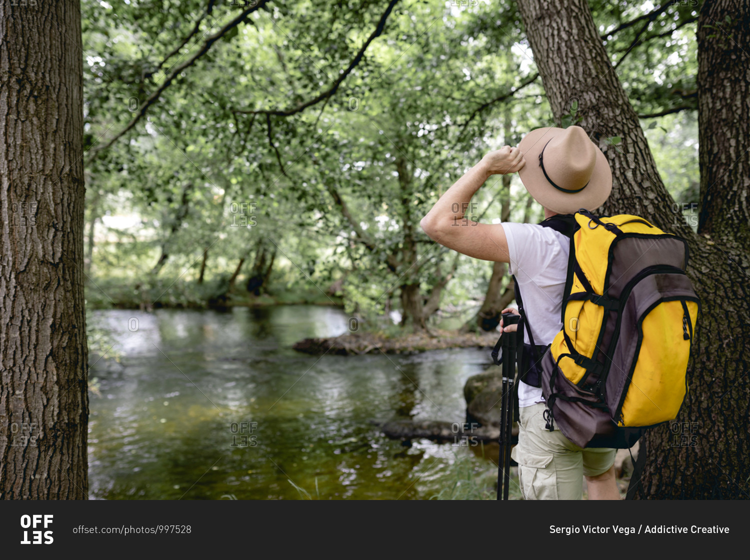young man doing a hiking trail with his yellow backpack and hat on his head by a lake with many trees and natural areas looking at the landscape