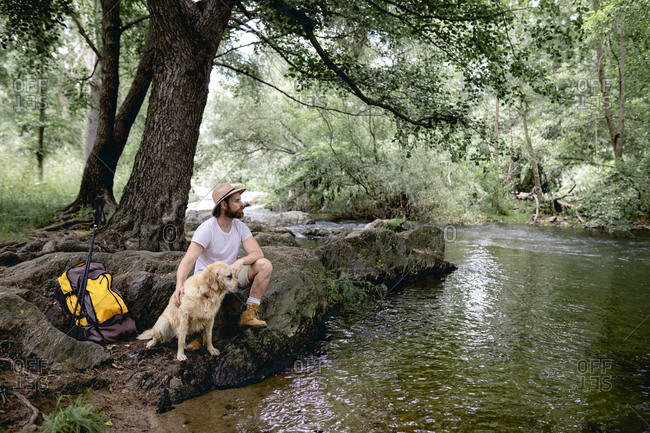 young man in a hat and yellow backpack sitting in the shade on the shore of a tree-filled lake lounging with his beige dog after hiking