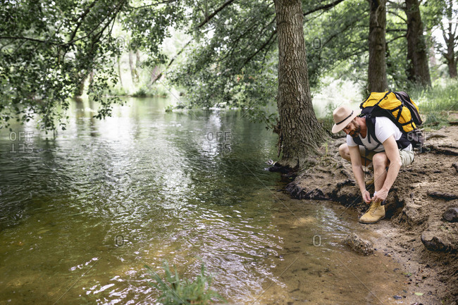 young man hiking with yellow backpack and hat tying his shoelaces on the shore of a tree-filled lake