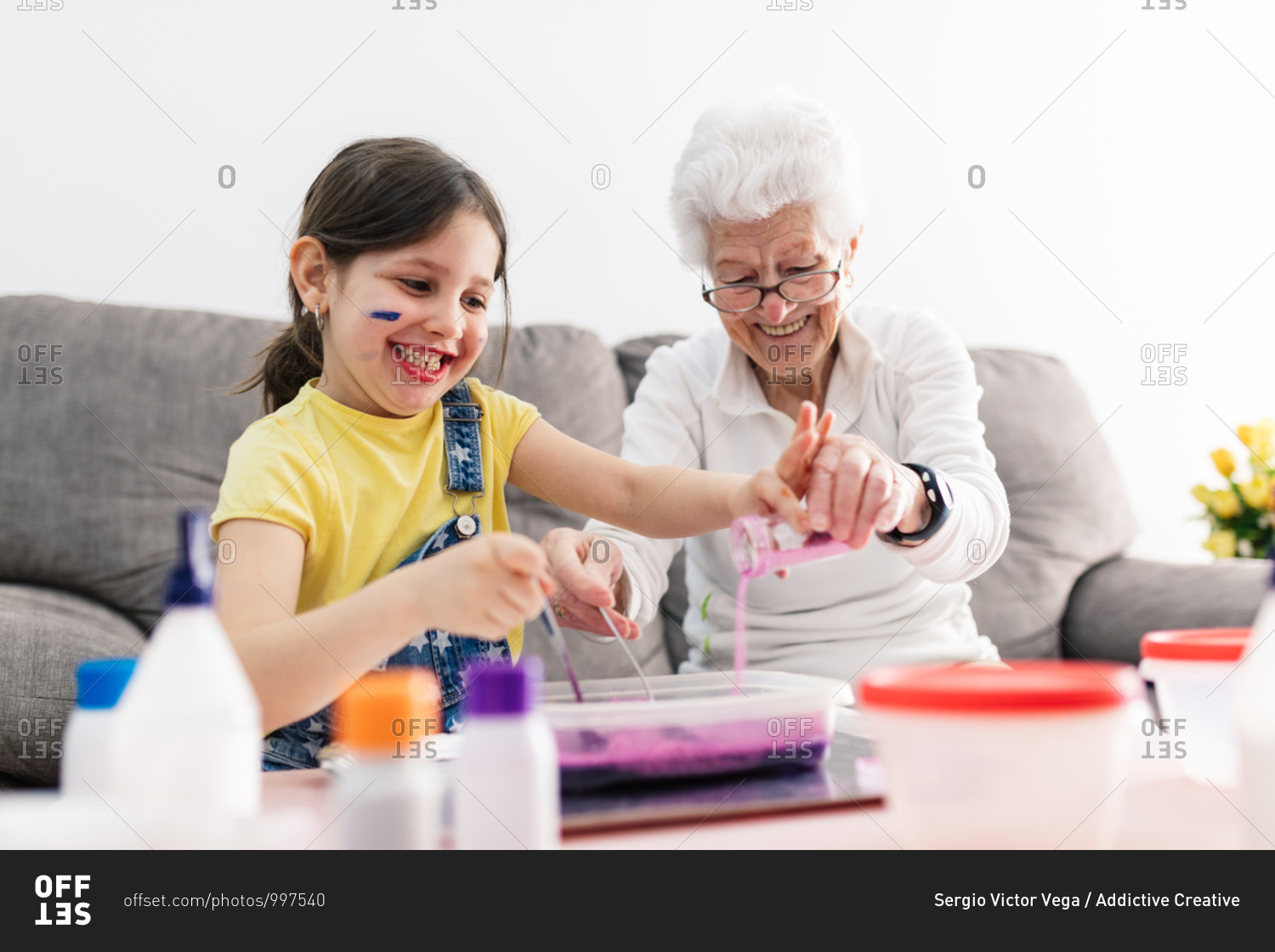 Smiling granny in eyeglasses helping positive granddaughter with dirty face in casual wear preparing blend using colorants in plastic container while sitting on comfortable couch in living room