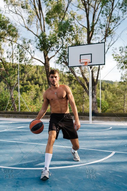 Concentrated male athlete with naked torso playing basketball alone on sports ground in summer