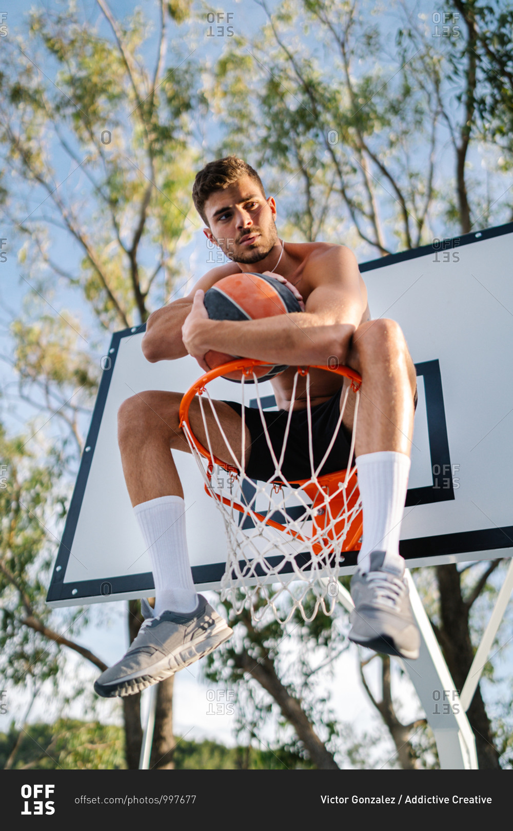 From below of male athlete in sportswear sitting on basketball hoop with ball and looking away