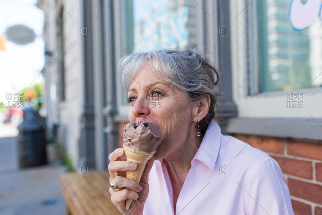 Senior woman sitting on sidewalk eating chocolate ice cream cone