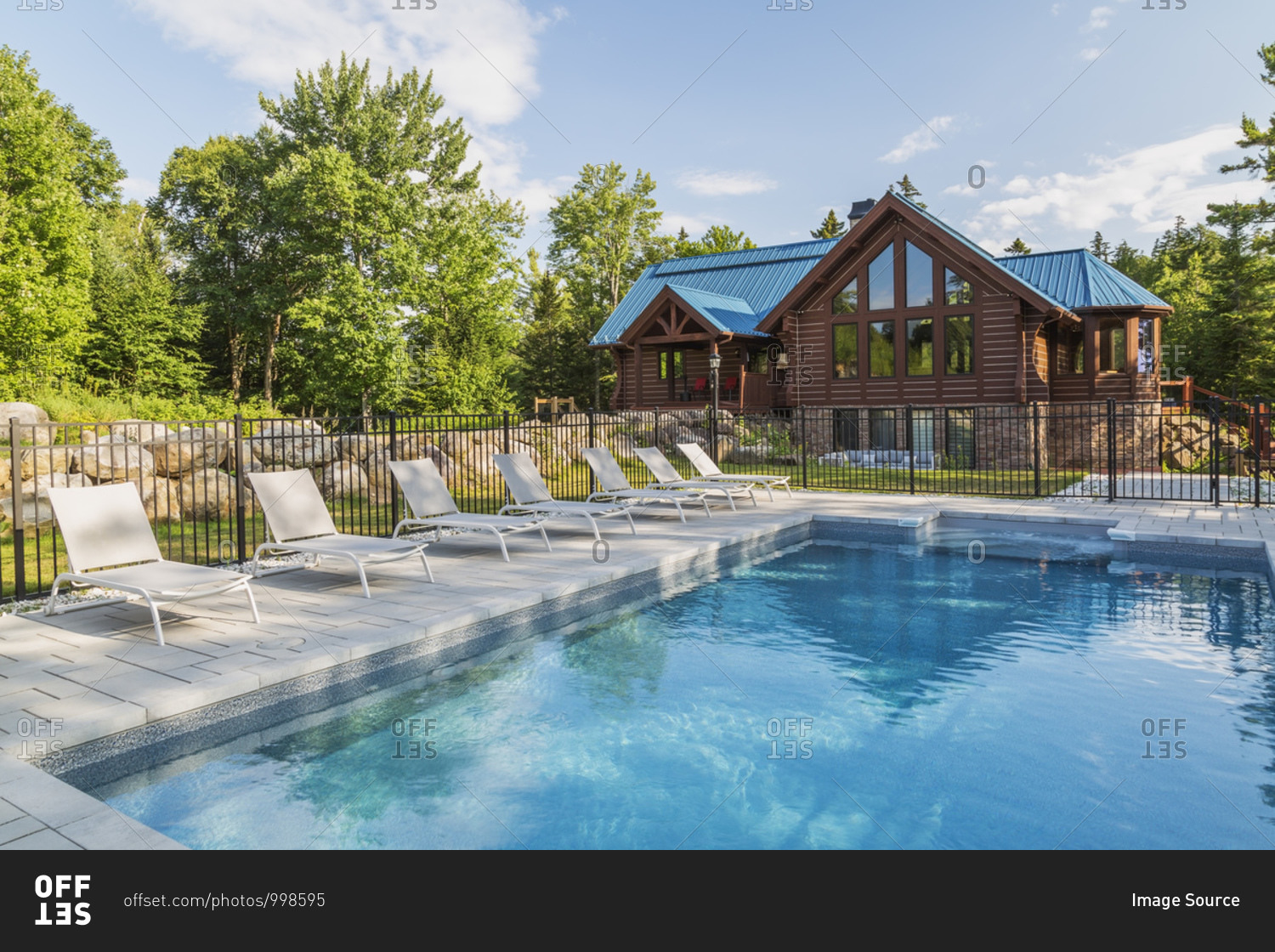 In-ground salt water swimming pool and brown stained milled Eastern white pine timber and flat log profile home facade with stone cladding on walk-out lower level and blue standing-seam sheet metal roof, Quebec, Canada