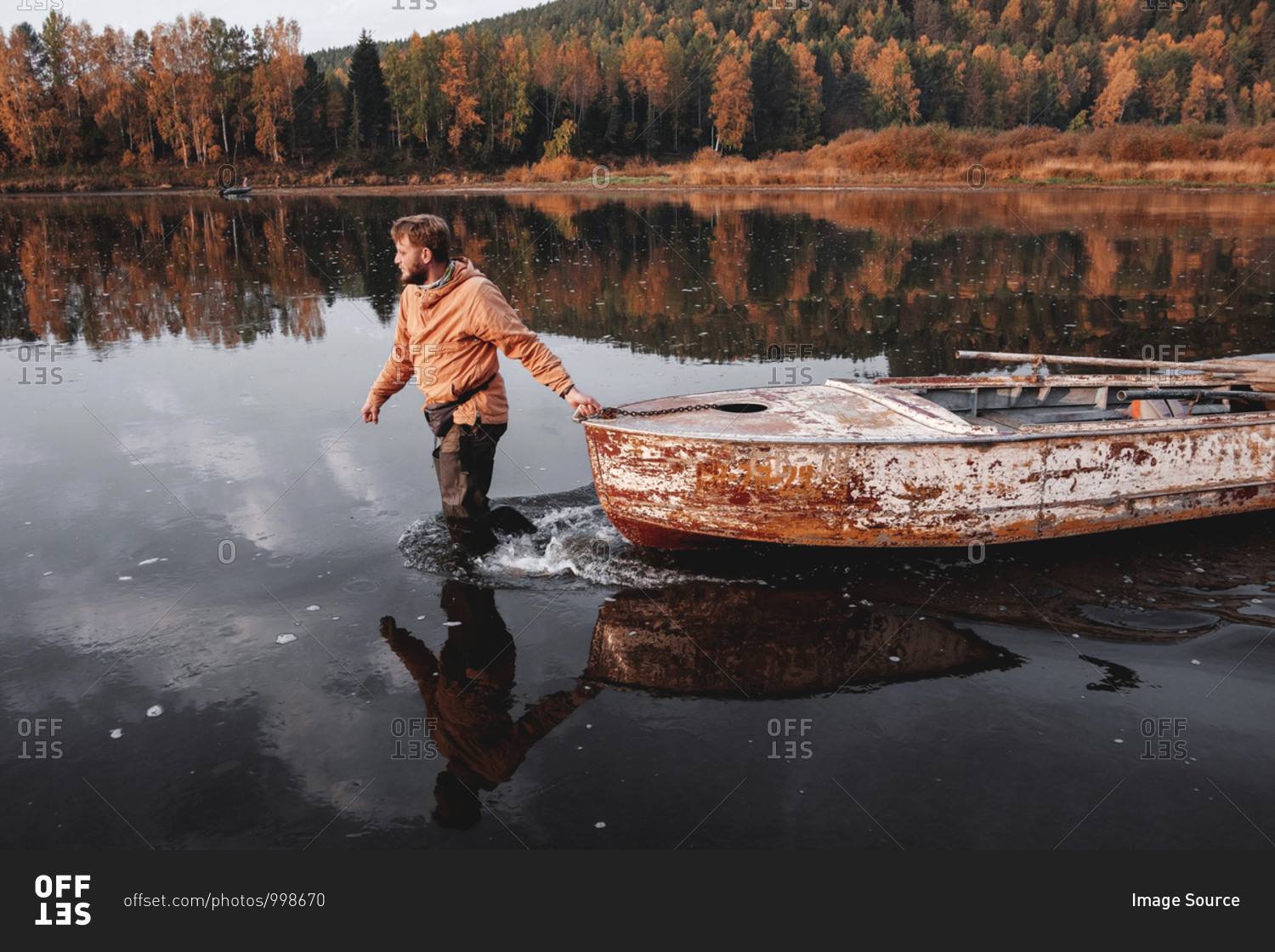 Man dragging old rusty boat in middle of lake in autumn, Russia