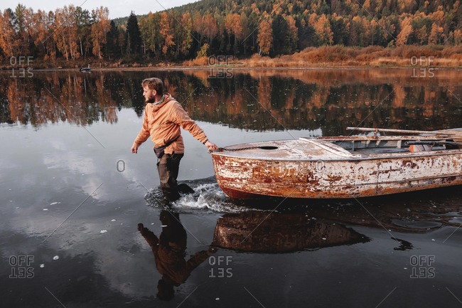 Man dragging old rusty boat in middle of lake in autumn, Russia