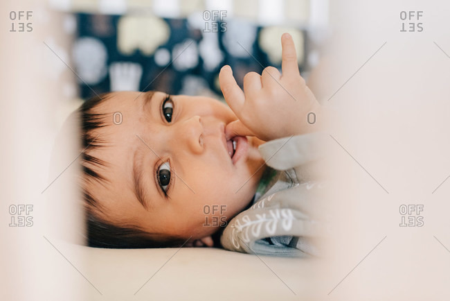 Baby boy lying awake and sucking thumb in crib, close up portrait