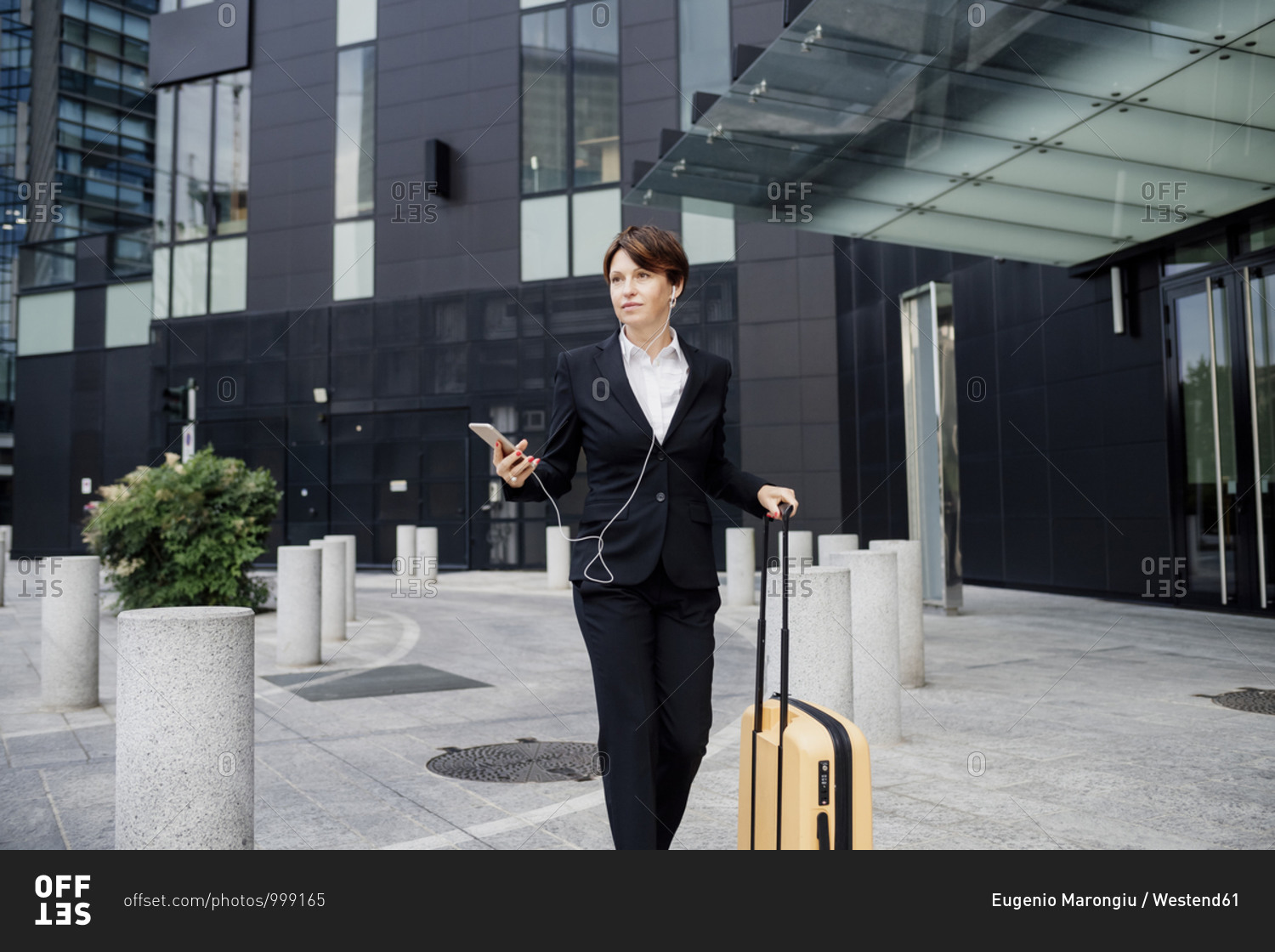 Female professional listening music while walking with suitcase against modern building