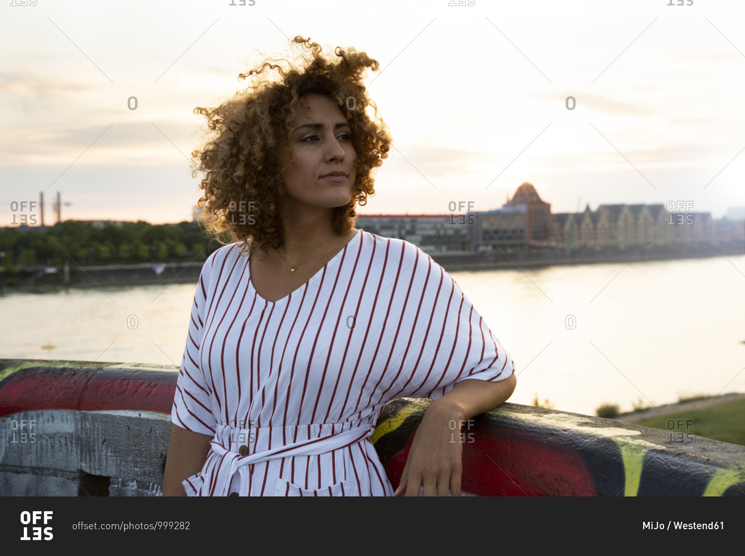 Thoughtful woman with curly hair standing by railing against river in city during sunset