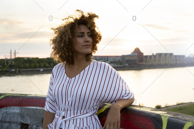 Thoughtful woman with curly hair standing by railing against river in city during sunset