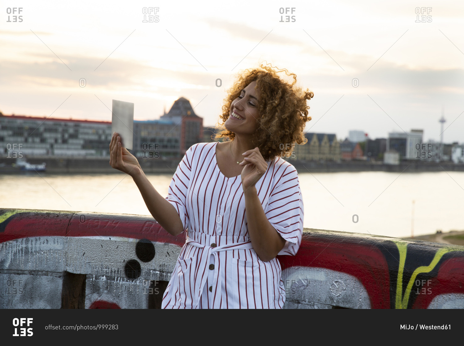 Smiling woman with curly hair holding digital tablet while standing against river in city at sunset