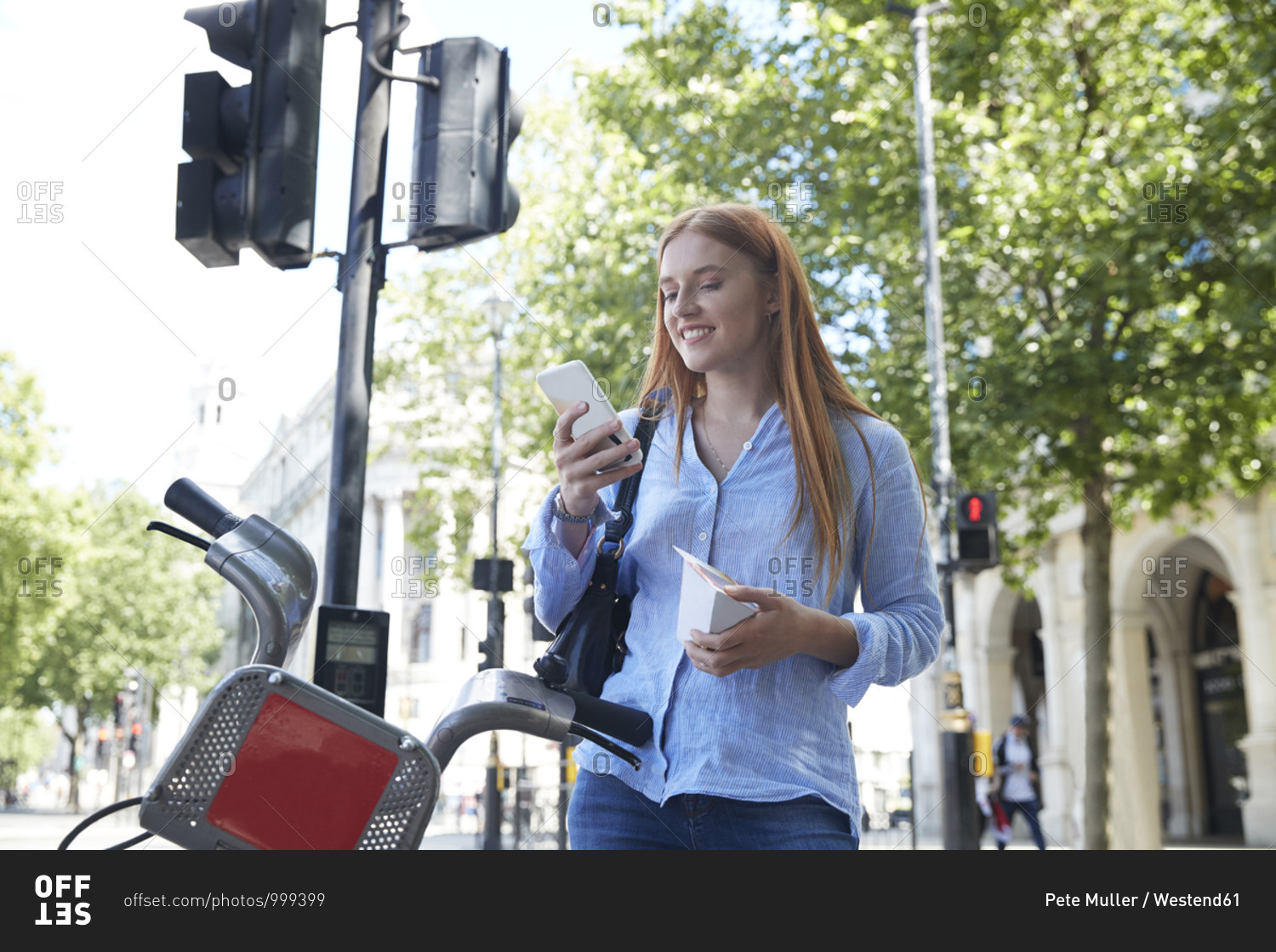 Happy young woman using smart phone while standing by hire bike in city