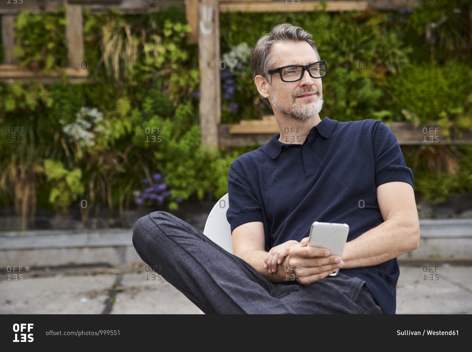 Thoughtful mature man holding smart phone while sitting on chair at yard